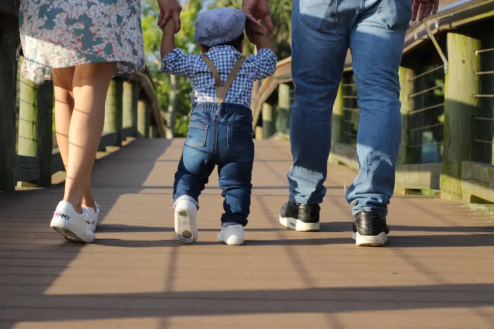 A toddler in suspenders walking between two adults on a wooden boardwalk